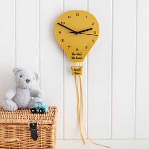 Puede incluir: Un reloj de globo aerostático amarillo con manecillas y números negros. El reloj tiene una cinta negra que cuelga de la parte inferior. El reloj está montado en una pared blanca. El texto en el reloj dice "The sky's the limit" y "Louise Anne".