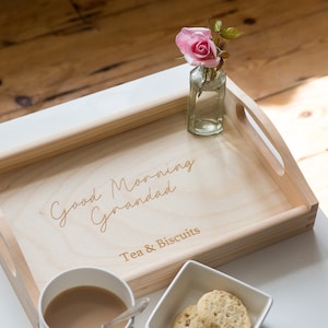 Può includere: Un vassoio di legno con le parole "Good Morning Grandad" e "Tea & Biscuits" incise su di esso. Il vassoio contiene una tazza di tè, due biscotti e un piccolo vaso con una rosa rosa.