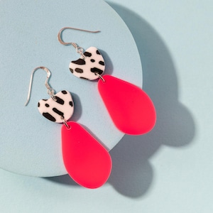 May include: A pair of dangle earrings. Each earring features a white heart-shaped top with black spots, connected to a bright pink teardrop-shaped bottom. The earrings hang from silver-colored hooks, set against a light blue background.