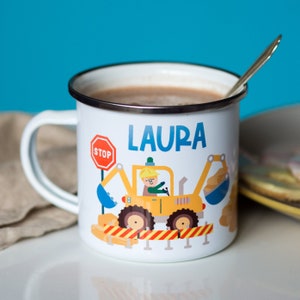 Op de afbeelding: Een witte emaille mok met een geel graafmachine-ontwerp, een stopteken en de naam "LAURA" erop gedrukt. De mok is gevuld met een bruine vloeistof en heeft een zilveren lepel op de rand.