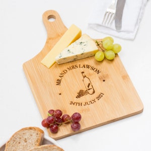 Può includere: Un tagliere per formaggi in bambù personalizzato con manico, inciso con "Mr. and Mrs. Lawson 24th July 2021" e un design con bottiglia di vino e uva.