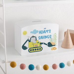 May include: White wooden bank with a curved top, featuring a yellow excavator illustration and the text "ADAM'S SAVINGS" in blue. The design includes a smiling sun, clouds, and trees. A slot is visible on top for inserting money.