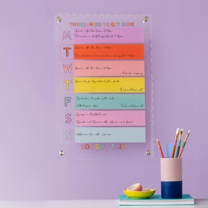 Puede incluir: Un planificador semanal de acrílico transparente con notas adhesivas de colores. Las notas tienen texto escrito a mano que incluye "Things I Need to Get Done", "Lunch with the fam", "Midweek shopping", "Dinner for dog walker by the plants", "Dad's turn to pick up the kids", "George is at football camp", "Drinks and Dinner with Annie and Lucas", "Afternoon tea with the group", y "You Got This!"