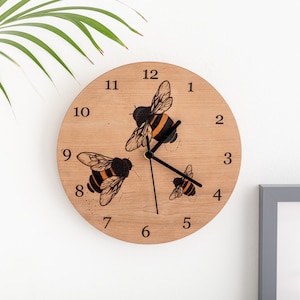 Könnte beinhalten: Eine Holzuhr mit einem Bienenmotiv. Das Zifferblatt der Uhr hat schwarze Zahlen und Zeiger, und drei Bienen sind in Schwarz und Orange dargestellt.