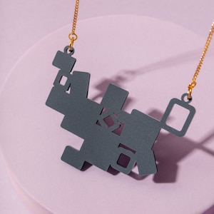 May include: A grey geometric pendant necklace with a gold chain. The pendant is made of multiple interlocking squares and rectangles.