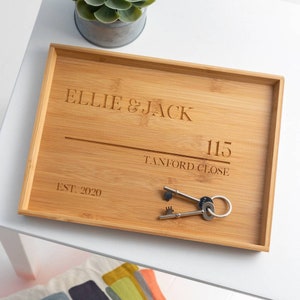 Può includere: Un vassoio in bambù personalizzato con il testo "Ellie & Jack", "115 Tanford Close" e "Est. 2020" inciso su di esso. Il vassoio è su una superficie bianca con un set di chiavi in cima.