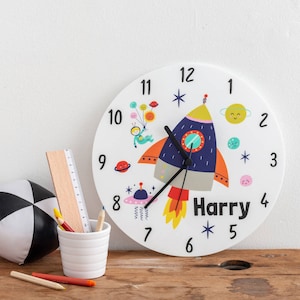 Peut inclure: Une horloge ronde blanche avec un motif de fusée. La fusée est bleue, orange et grise avec une flamme jaune. Le cadran de l'horloge a des chiffres et des aiguilles noirs. L'horloge a un fond blanc avec des étoiles, des planètes et un petit astronaute avec des ballons. Le nom "Harry" est écrit en lettres noires sous la fusée.
