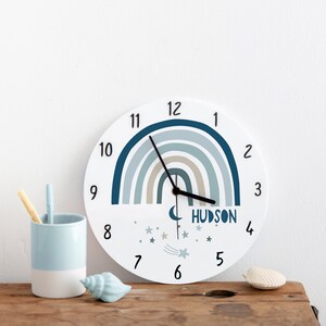 Może przedstawiać: Biały okrągły zegar z niebieskim i beżowym wzorem tęczy. Tarcza zegara ma czarne wskazówki i czarne cyfry. Zegar ma półksiężyc i gwiazdy z gwiazdą spadającą. Zegar ma nadrukowane imię "HUDSON".