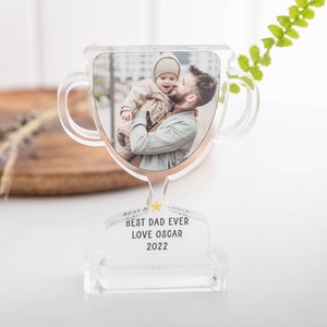 May include: Clear acrylic trophy with a photo of a man holding a baby. The trophy reads "BEST DAD EVER LOVE OSCAR 2022". The trophy has a gold-coloured trim and a star accent.