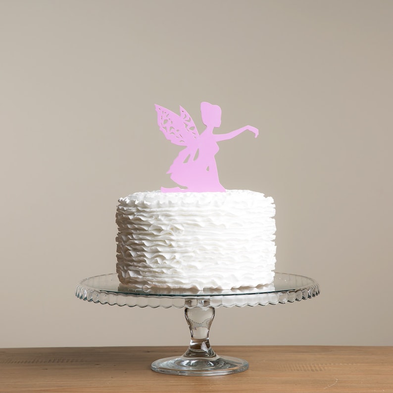 May include: A white cake with ruffled frosting topped with a pink paper cutout of a fairy.