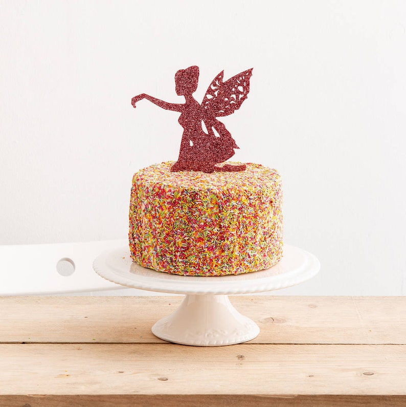 May include: A white cake decorated with colorful sprinkles and topped with a pink glitter fairy cake topper.