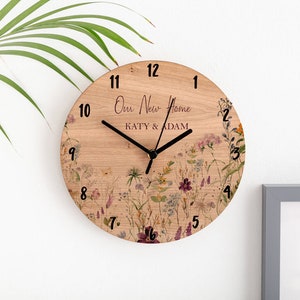 Puede incluir: Un reloj de madera redondo con un diseño floral y agujas negras. La esfera del reloj tiene los números del 1 al 12 y el texto "Our New Home KATY & ADAM".