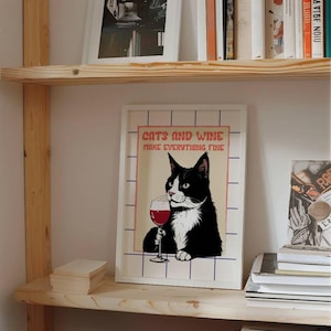 Puede incluir: Un cuadro enmarcado con un gato blanco y negro sosteniendo una copa de vino tinto. El cuadro tiene el texto "CATS AND WINE MAKE EVERYTHING FINE" en un estilo retro. La obra de arte se exhibe en una estantería de madera.