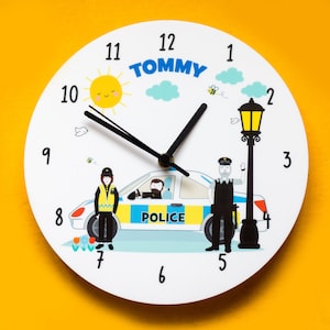 May include: A white clock with black hands and a blue and yellow police car design. The clock face has numbers 1 through 12, and the words "TOMMY" are printed at the top. The clock is surrounded by a yellow background.