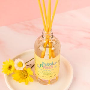 May include: A glass reed diffuser with yellow reeds and a yellow label reading "Petal & Musk Mini Reed Diffuser". The diffuser is on a white plate with yellow flowers. The background is pink.
