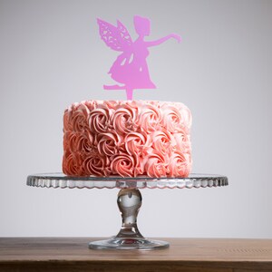May include: A pink frosted cake decorated with pink frosting swirls, topped with a pink fairy silhouette cake topper. The cake is on a glass cake stand.