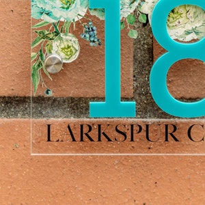 May include: Clear acrylic house number sign with the number 18 in turquoise. The sign features a floral design with white and teal flowers and the text "Larkspur C" in black.