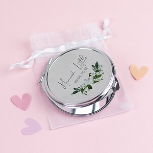 May include: A silver compact mirror with a floral design and the text "Hannah Little Bride to Be" on the front. The mirror is in a white organza bag.