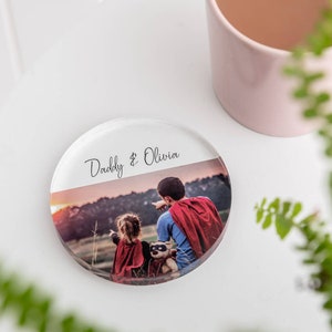 Puede incluir: Un posavasos redondo de vidrio con una foto de un padre y una hija vestidos de superhéroes. El texto "Daddy & Olivia" está impreso en el posavasos.