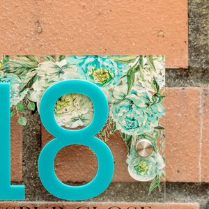 May include: Clear acrylic house number sign with a turquoise number 8 and floral design. The sign features a turquoise number 8 and a floral pattern with teal and white flowers. The sign is mounted on a brick wall.