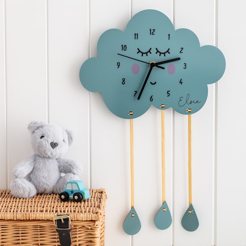 May include: A blue cloud-shaped clock with a black face and white numbers. The clock has a pair of eyelashes and a smiling mouth. The clock is hanging from the ceiling with three gold strings that end in blue raindrops. The clock is personalized with the name "Elodie".
