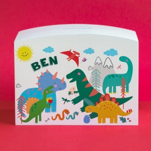 Könnte beinhalten: Eine weiße Holz-Dinosaurier-Spardose mit einem Schlitz oben. Die Dose ist mit einer bunten Illustration von Dinosauriern, einer Sonne und dem Namen "BEN" verziert.