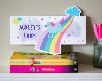 May include: A white wooden door sign with a rainbow, a unicorn, and stars. The sign says "Audrey's Room" in purple letters.