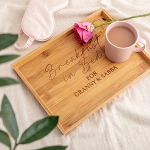 Può includere: Un vassoio di legno per la colazione a letto con la scritta "Breakfast in Bed for Granny & Sabba" incisa su di esso. Un rosa rosa e una tazza rosa con caffè sono sul vassoio.