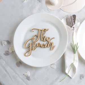 Puede incluir: Un plato blanco con un letrero de madera que dice "The Groom" en letra cursiva. El plato está sobre un mantel blanco con confeti blanco y gris esparcido.