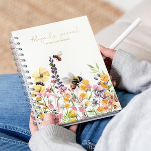 Può includere: Un diario a spirale bianco con le parole "Abigail's Journal" e "Notes and doodles" stampate in lettere dorate. La copertina presenta un'illustrazione ad acquerello di un campo di fiori con api.