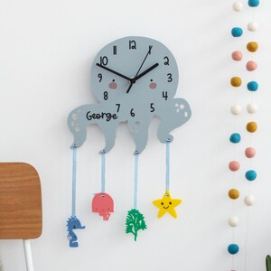 May include: A gray octopus-shaped clock with a white face and black hands. The clock has a blue ribbon hanging from each tentacle, with a seahorse, a starfish, a mushroom, and a seaweed charm attached. The clock is personalized with the name "George".