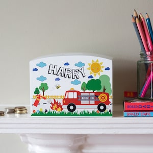 Op de afbeelding: Witte houten spaarpot met een rode brandweerwagen, een gele zon, groene bomen en blauwe wolken. De naam "HARRY" staat in het zwart op de voorkant.