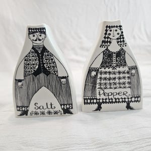 May include: Two white ceramic salt and pepper shakers with black ink illustrations. The salt shaker features a figure in a vest and striped trousers, while the pepper shaker depicts a figure in a patterned dress. Both have the words Salt and Pepper.