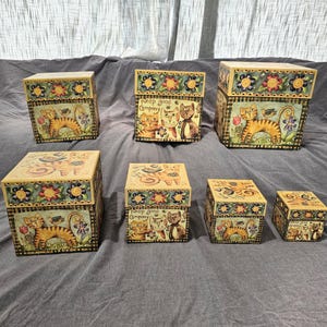 Puede incluir: Un conjunto de seis cajas decorativas cuadradas de varios tamaños, con ilustraciones pintadas de gatos atigrados naranjas, flores y pájaros. Algunas cajas muestran el texto 'Keep good company' o 'Shine your light' con un borde a cuadros amarillo y negro.