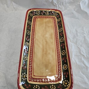 Puede incluir: Una bandeja rectangular de cerámica con un centro de color tostado liso bordeado por un borde verde oscuro. El borde está decorado con un patrón repetitivo de pequeñas flores blancas y amarillas y formas de hojas rojas, con un borde exterior rojo.