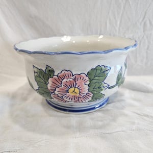 Puede incluir: Un cuenco de cerámica blanca con borde festoneado y ribete pintado en azul. El exterior presenta un diseño floral pintado a mano con una flor grande de color rosa, centro amarillo y hojas verdes. La base está decorada con bandas circulares azules.