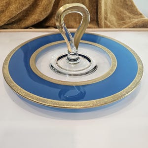 May include: A round glass serving tray featuring a clear centre, a wide blue border, and a gold-toned rim. A central clear glass loop handle rises from the middle of the tray, providing a grip for carrying the circular dish with its decorative gold trim.
