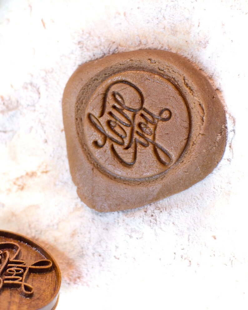 May include: A close-up of a brown cookie dough with the words "Holly Jolly" stamped into the dough. The dough is on a white surface covered in powdered sugar.