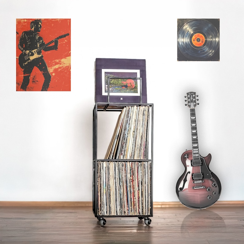 May include: A black metal record storage unit with four shelves filled with vinyl records. A black electric guitar with a red-brown finish is leaning against the unit. A black and white poster of a person playing a guitar is hanging on the wall behind the unit. A black and white record with a red center is hanging on the wall to the right of the unit.