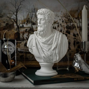 May include: White plaster bust of a man with a beard, wearing a toga-like garment. The bust is on a white pedestal and is in front of a painting.