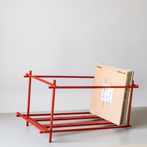 Puede incluir: Un soporte de discos de metal rojo con un marco rectangular. El soporte está diseñado para exhibir discos de vinilo. Se ve una pila de discos, con la portada del álbum "Primitive Man" hacia adelante.