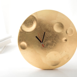 May include: A gold-colored clock with a round face and a textured surface. The clock has black hands and a red second hand.