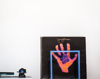 Könnte beinhalten: Eine schwarze Schallplatte mit einem Handabdruck-Design und dem Text "George Harrison" auf dem Cover. Die Schallplatte wird in einem blauen Metallständer präsentiert.