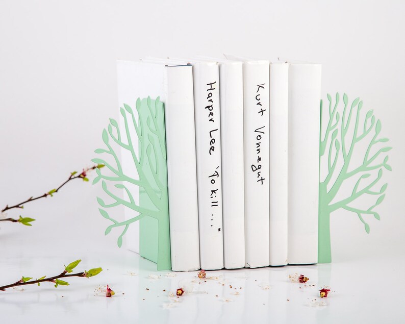 May include: A pair of green metal bookends shaped like trees holding up five white books. The books have the titles "Harper Lee 'To Kill..." and "Kurt Vonnegut" written on them.