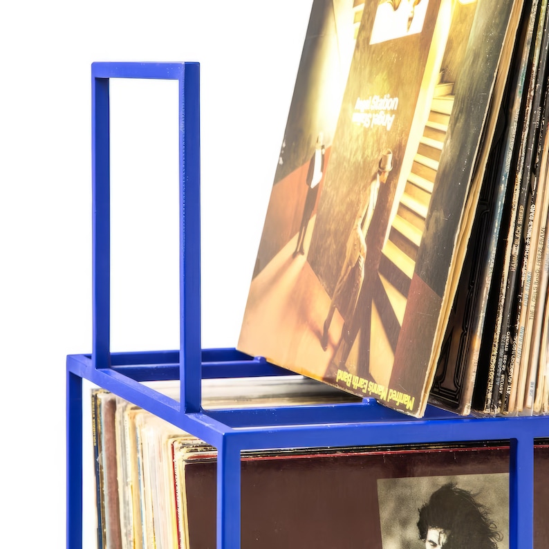 May include: A blue metal record storage rack with a handle. The rack is filled with vinyl records, including one with the album cover "Angel Station" by "Howard Jones".