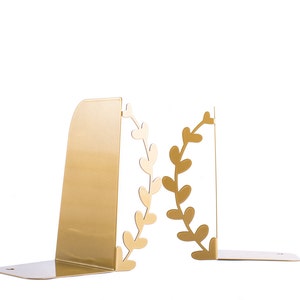 May include: Gold metal bookends with a leafy design. The bookends are shaped like a wreath with leaves and are a decorative way to hold books upright.
