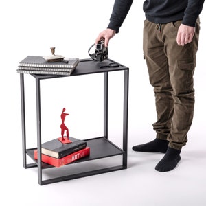 May include: A black metal side table with a minimalist design. The table has two shelves and a rectangular frame. On the top shelf are notebooks, a decorative object, and a camera. The lower shelf holds books and a red sculpture.
