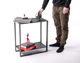 May include: A black metal side table with a minimalist design. The table has two shelves and a rectangular frame. On the top shelf are notebooks, a decorative object, and a camera. The lower shelf holds books and a red sculpture.