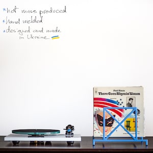 May include: A white turntable with a black platter and a blue record player needle. A blue metal record stand holds a vinyl record with the title "There Goes Rhymin' Simon" by Paul Simon. The white wall behind the turntable has handwritten text: "*hot mass produced *hand welded *designed and made in Ukraine"