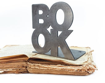 May include: A metal bookend shaped like the word "BOOK" in all caps. The bookend is sitting on top of an old, worn book.